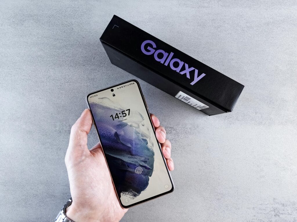 Close-up of a hand holding a smartphone with a Galaxy box on a neutral background.