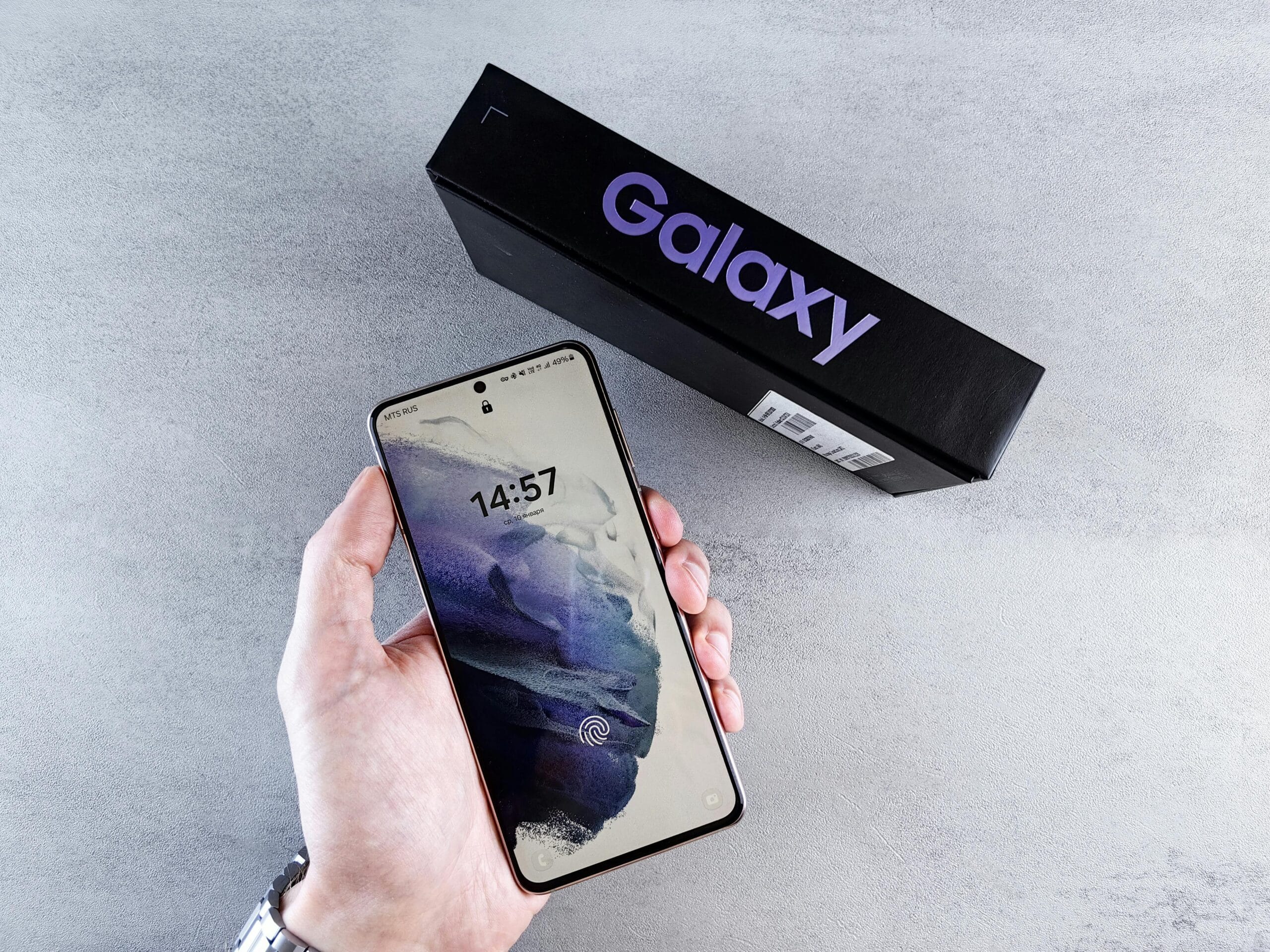 Close-up of a hand holding a smartphone with a Galaxy box on a neutral background.